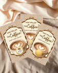 Three identical books titled "Who Spilled the Coffee?" with illustrations of coffee cups and hearts on the covers, placed on a beige fabric background.